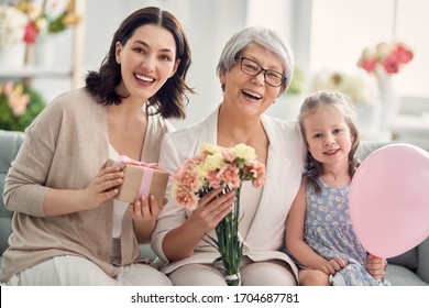 Happy Mother's Day! Child Daughter Is Congratulating Mom And Granny Giving Them Flowers And Gift. Grandma, Mum And Girl Smiling And Hugging. Family Holiday And Togetherness. 