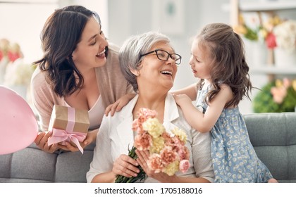 Happy Mother's Day! Child Daughter Is Congratulating Mom And Granny Giving Them Flowers And Gift. Grandma, Mum And Girl Smiling And Hugging. Family Holiday And Togetherness. 