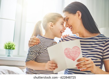 Happy mother's day! Child daughter congratulates mom and gives her postcard. Mum and girl smiling and hugging. Family holiday and togetherness.