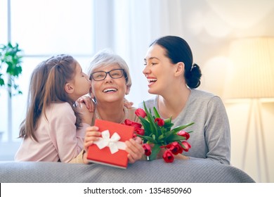 Happy Mother's Day! Child And Mom Congratulating  Granny Giving Her Flowers Tulips Ang Gift Box. Grandma, Mum And Girl Smiling And Hugging. Family Holiday And Togetherness.