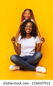 Happy Mother's Day Concept. Beautiful African American Mom Holding Hands With Her Cute Daughter, Sitting On Floor Over Yellow Studio Background With Copy Space