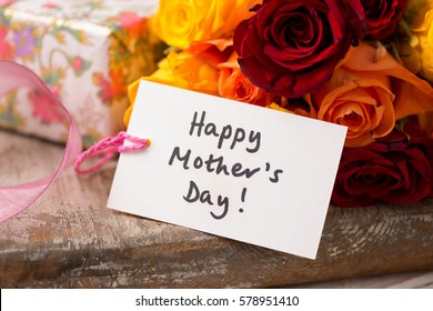 A "Happy Mother's Day" gift card or tag in front of a bouquet of roses and a wrapped present on a wooden surface