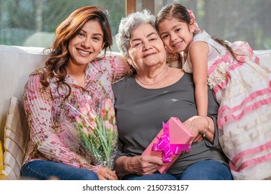 Happy Mother's Day! Latin Girl Mom And Grandmother Giving Flowers And Gifts. Grandma, Mom, And Little Girl Smiling And Hugging.