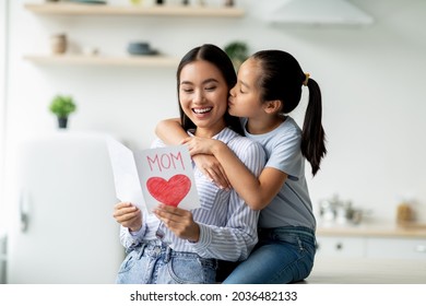 Happy Mother's Day. Little Girl Greeting Her Mom With Gift Card With Big Red Heart And Kissing Mommy In Cheek, Sitting Together In Kitchen Interior, Copy Space