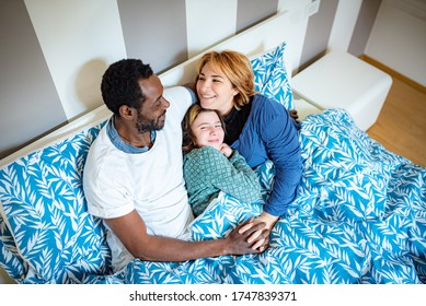 Happy Multi Ethnic Family Hugs In Bed As Soon As They Wake Up, Caucasian Mom And African Dad Hug Their Daughter Tenderly, Cuddles In Bed In The Morning As Soon As They Wake Up