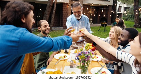 Happy Multi Ethnic Family Sitting At Table With Meal Outdoor At Picnic And Toasting With Juice And Wine. Joyful Young And Old People Having Dinner And Toast At Party Barbrque. Celebration On Weekend.