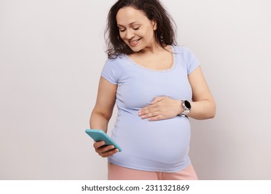 Happy multi ethnic young pregnant woman holding her belly, feeling baby movements, smiling, checking pregnancy mobile application on his smartphone, isolated over white background. People. Technology - Shutterstock ID 2311321869