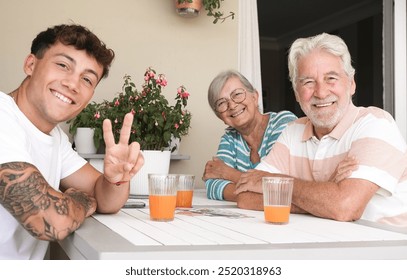 Happy multi generation family of young handsome grandson and white haired grandparents enjoying free time together sitting outdoors on terrace smiling drinking an orange juice, looking at camera - Powered by Shutterstock