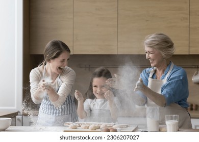 Happy multi generational women have fun while prepare homemade pastries, preschooler girl her mature grandma and young mother clap their hands, throwing flour, making dough for pie for family holiday - Powered by Shutterstock