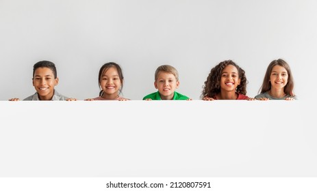 Happy Multicultural Children Posing With Empty White Poster Looking At Camera Over Gray Studio Background. Boys And Girls Advertising Your Text Posing Over Blank Board. Panorama, Mockup - Powered by Shutterstock