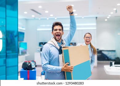 Happy Multicultural Couple Dressed Elegant Holding Box With New Television In It. Tech Store Interior.