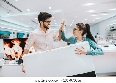 Happy Multicultural Couple Holding New Smart Tv In Box And Giving High Five While Standing At Tech Store.