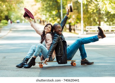 Happy multicultural couple sitting on a longboard on a street and rising hands with hats 