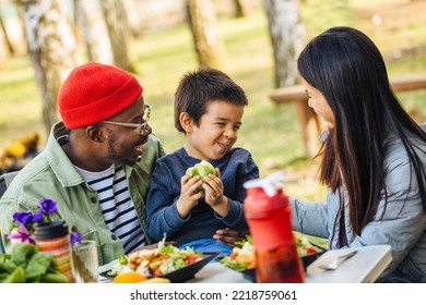 A Happy Multicultural Family Eats Healthy Food In Nature In Front Of The Van. A Man Holds A Boy Eating The Apple And Talks To Him While His Mother Smiles At Them.