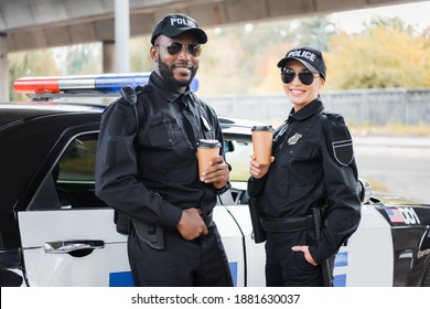 happy multicultural police officers with paper cups looking at camera near patrol car on blurred background on urban street