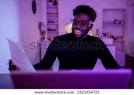 A happy multicultural statistician is sitting at home late at night in a purple lighted room and working on paperwork. A smiling data analyst is working on a project while smiling at paperwork at home
