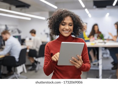 Happy multiethnic businesswoman with curly hair using digital tablet in office. Successful casual mixed race business woman working on laptop in workplace with colleagues sitting at desk in background - Powered by Shutterstock