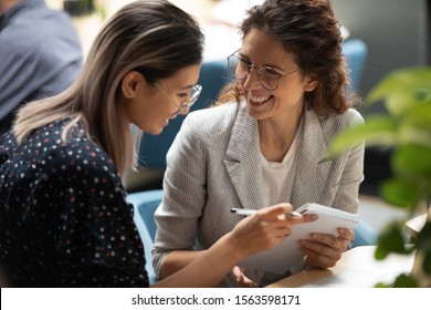 Happy multiethnic colleagues sitting together at coworking place, discussing notes or insights from educational training workshop. Smiling caucasian businesswoman sharing ideas with asian teammate.