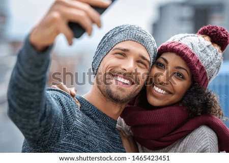 Happy multiethnic couple in the city taking photo in a winter day. Smiling man and african girl taking selfie with smartphone on street. Young woman and cheerful guy on christmas holidays.