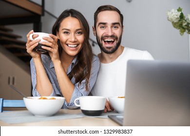 Happy multiethnic couple having breakfast at the kitchen, looking at laptop computer స్టాక్ ఫోటో