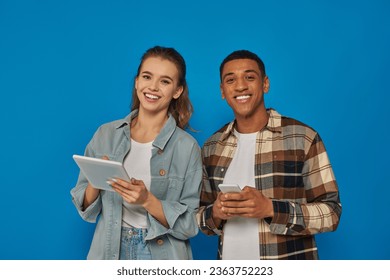 happy multiethnic couple holding smartphone and digital tablet on blue backdrop, look at camera - Shutterstock ID 2363752223
