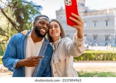 Happy multi-ethnic couple taking selfie and smiling in a park with city views - Powered by Shutterstock