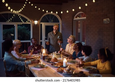 Happy Multiethnic Extended Family Gathering For Dinner At Dining Table On A Patio. Focus Is On Happy Man Holding A Toast. 