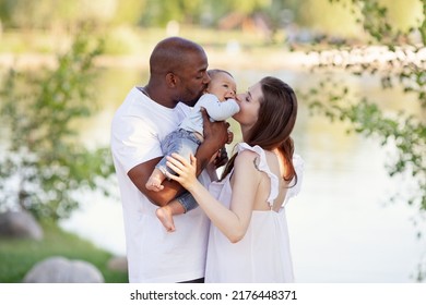 Happy Multiethnic Family. Caucasian Mother And African American Father Kiss Small Child. Parents, Portrait Of Mom, Dad And Baby On Hands Over Nature Background