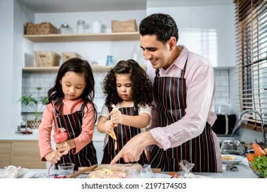 Happy Multiethnic Family Cooking Creams Cake Together In The Kitchen At Home. A Father Teaches Daughters To Make Homemade Desserts, Enjoy Leisure Activities Time