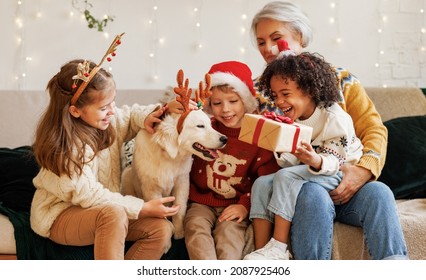 Happy multiethnic family with dog on Christmas day, little kids siblings of different races spend winter holidays at grandma house. Loving senior grandmother receiving New Year gift from grandchildren