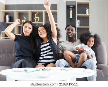 A Happy Multi-ethnic Family Sitting On The Sofa With A Cute Daughter Raising Her Hand In Congratulations. Couple Smiling And Looking At The Camera Asian Mom And Black Dad Relax Together At Home