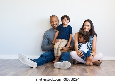 Happy multiethnic family sitting on floor with children. Smiling couple sitting with two sons and looking at camera. Mother and black father with their children leaning on wall with copy space. - Powered by Shutterstock