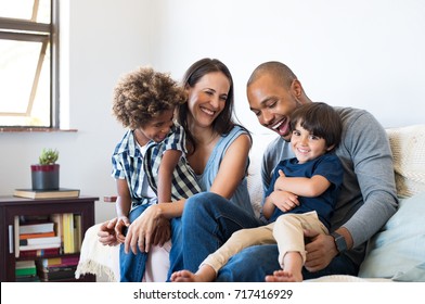 Happy multiethnic family sitting on sofa laughing together. Cheerful parents playing with their sons at home. Black father tickles his little boy while the mother and the brother smile. - Powered by Shutterstock