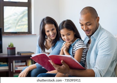 Happy Multiethnic Family Sitting On Sofa Together Looking At Daughter's Drawing Book. Black Father Reading The Homework On Exercise Book Of His Cute Kid. Parents Feeling Proud On Progress Of Daughter.