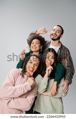 happy multiethnic friends in stylish outfit posing with sweet lollipops isolated on grey