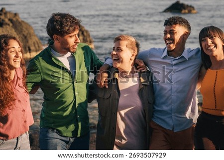 A happy multi-ethnic group of five young friends in casual clothing are walking arm-in-arm on a rocky beach, laughing and bonding together during a summer vacation.