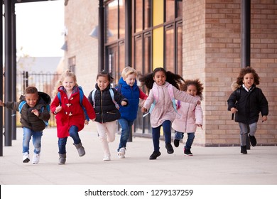 A Happy Multi-ethnic Group Of Young School Kids Wearing Coats And Carrying Schoolbags Running In A Walkway With Their Classmates Outside Their Infant School Building