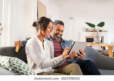 Happy multi-ethnische geschlechtsreife Ehepaare mit digitalem Tablet für Online-Bezahlung mit Kreditkarte. Lateinische Ehefrau, die ihrem indischen Ehemann etwas zum Kauf auf digitalem Tablet zeigt. Nahost Paar entspannend.