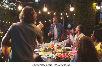 Happy multi-ethnic people having fun at garden at night. Caucasian boys and girls drinking and eating while talking outdoor. African American female laughing. Asian girl with watermelon. Party concept - Powered by Shutterstock