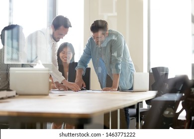 Happy multiethnic team working together brainstorm on paperwork gather at office table briefing, diverse workers group discussing project results in teamwork at corporate meeting, behind glass view