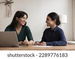 Happy multiethnic women have fun seated at desk in workplace. Female teammates, distracted from work, tell jokes, laughing, having goods friendly work relations, engaged in teamwork and communication