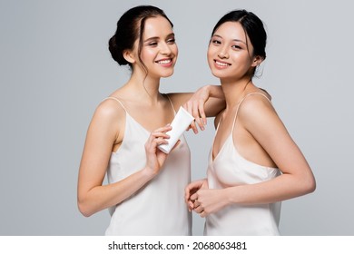 Happy multiethnic women in satin dresses holding cream in tube isolated on grey