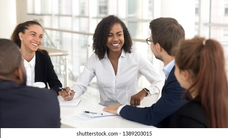 Happy multiethnic work group gather in conference room discuss share ideas talk at company meeting, smiling diverse employees speak considering startup brainstorming together at team briefing