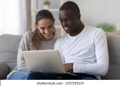 Happy Multiethnic Young Couple Sit Relax On Couch At Home Using Laptop Shopping Online Together, Smiling International Husband And Wife Rest In Living Room Watch Movie On Computer, Browse Internet