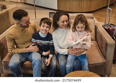 Happy Multi-ethnic Young Loving Mom And Dad Spending Time Together Playing Toy Planes With Their Kids, Son And Daughter At The Airport While Waiting For A Flight. Family Air Travel Concept