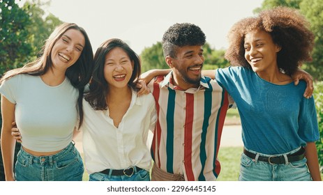Happy multiethnic young people walk embracing on summer day outdoors. Group of friends are talking and laughing merrily while walking along path in city park