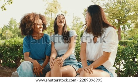 Happy multiethnic young women talking while sitting on park bench on summer day outdoors. Group of girls talking and laughing merrily in city park