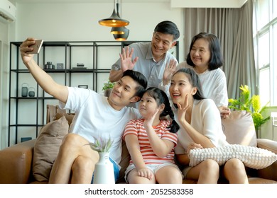 Happy multi-generation Asian family with kid daughter on sofa taking selfie with smartphone and having fun together at home.