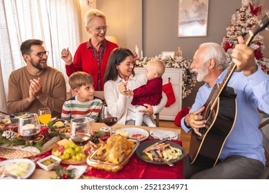 Happy multi-generation family gathered around the table, senior man playing the guitar and singing Christmas songs while having Christmas dinner all together at home - Powered by Shutterstock