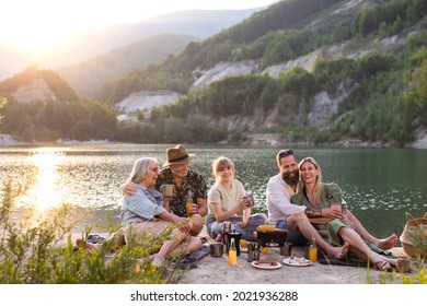 Happy Multigeneration Family On Summer Holiday Trip, Barbecue By Lake At Sunset.